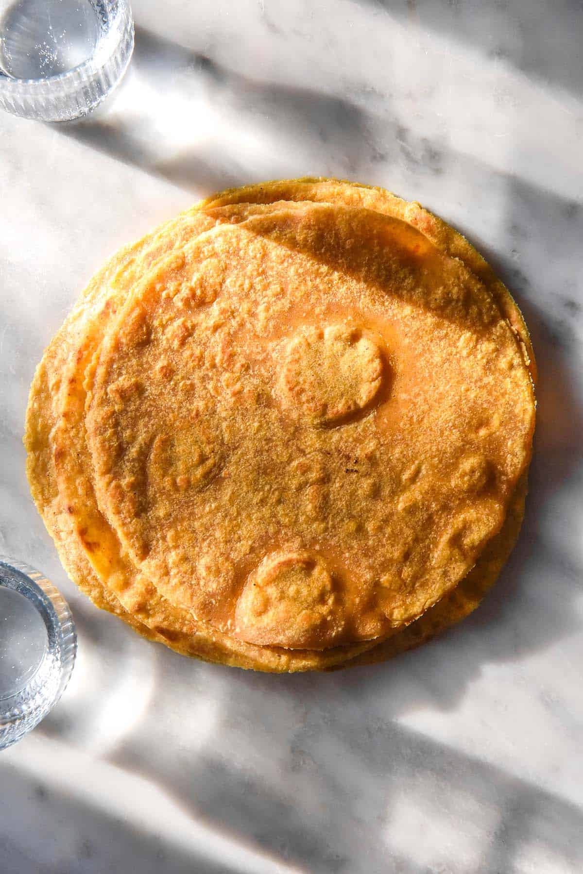A brightly lit aerial image of a stack of gluten free sweet potato tortillas on a white marble table. Glasses of water cast shadows to the top and bottom of the tortillas