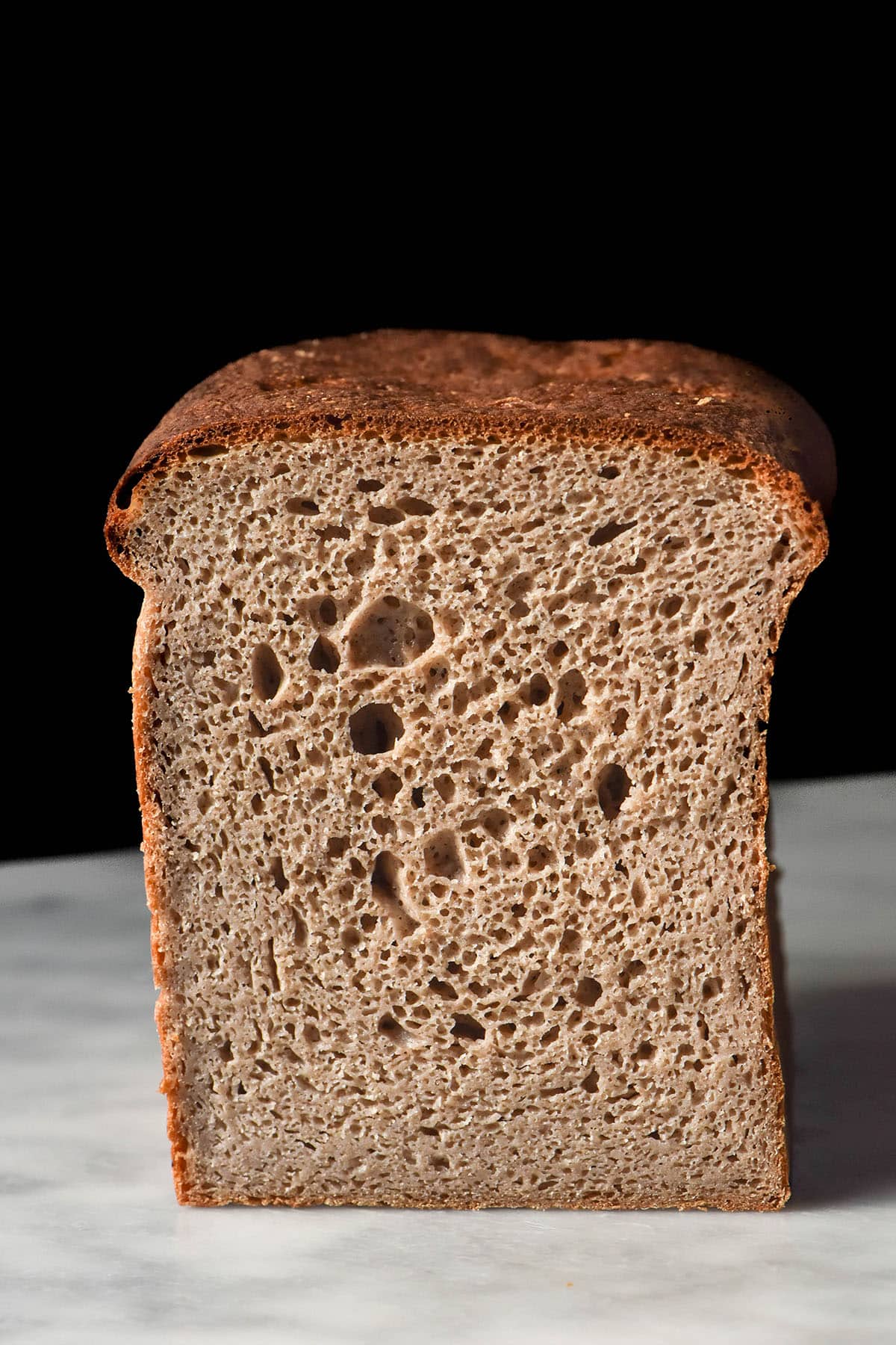 A side on macro image of a loaf of gluten free buckwheat sourdough on a white marble table against a black backdrop. The bread has been sliced to reveal the inner crumb.