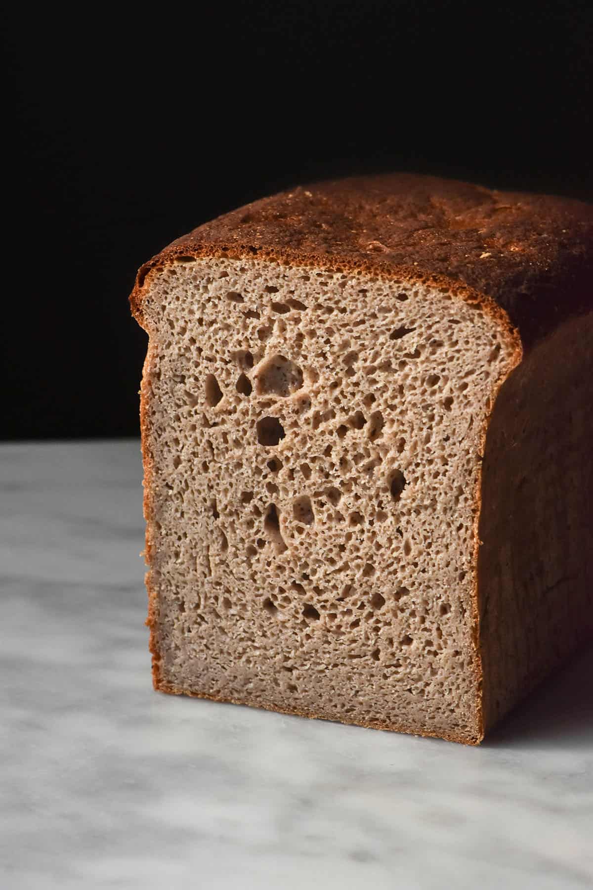 A close up side on image of a loaf of gluten free buckwheat sourdough bread. The loaf sits on a white marble table against a dark backdrop. It has been sliced to reveal the open crumb of the bread.