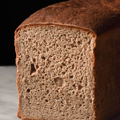 A close up side on image of a loaf of gluten free buckwheat sourdough bread. The loaf sits on a white marble table against a dark backdrop. It has been sliced to reveal the open crumb of the bread.