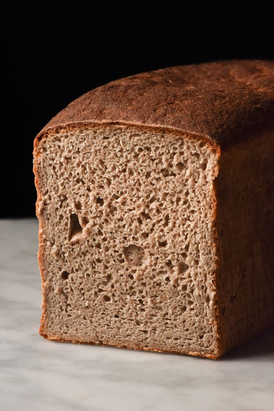 A close up side on image of a loaf of gluten free buckwheat sourdough bread. The loaf sits on a white marble table against a dark backdrop. It has been sliced to reveal the open crumb of the bread.