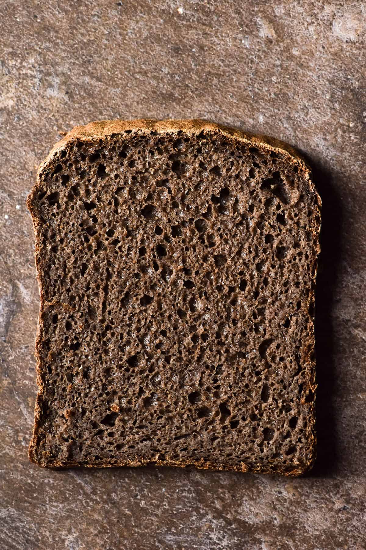 An aerial macro image of a slice of gluten free dark buckwheat bread on a medium brown backdrop. 