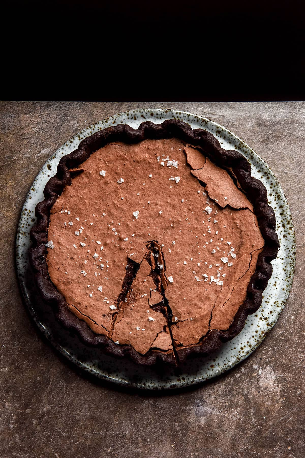 A moody aerial image of a gluten free brownie pie with a chocolate crust. The pie is topped with sea salt flakes and sits on a white speckled ceramic plate atop a brown backdrop