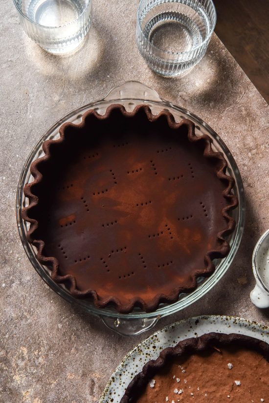 An aerial image of a gluten free chocolate pastry pie crust that is crimped into a pie plate and sitting atop a brown backdrop. Water glasses, a bowl of salt and a gluten free brownie pie are casually arranged around the pie crust.