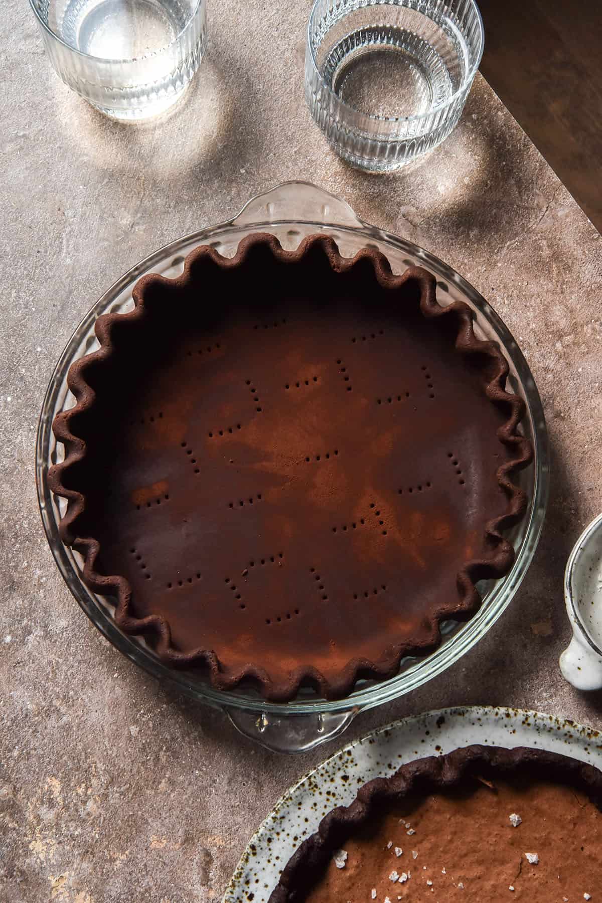 An aerial image of a gluten free chocolate pastry pie crust that is crimped into a pie plate and sitting atop a brown backdrop. Water glasses, a bowl of salt and a gluten free brownie pie are casually arranged around the pie crust.