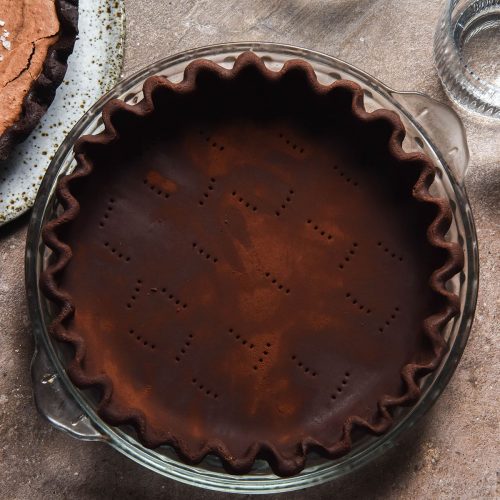 An aerial image of gluten free chocolate pastry pressed and crimped into a pie plate atop a light brown backdrop