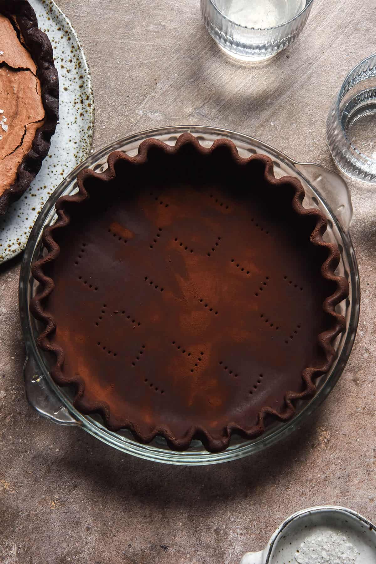 An aerial image of gluten free chocolate pastry pressed and crimped into a pie plate atop a light brown backdrop
