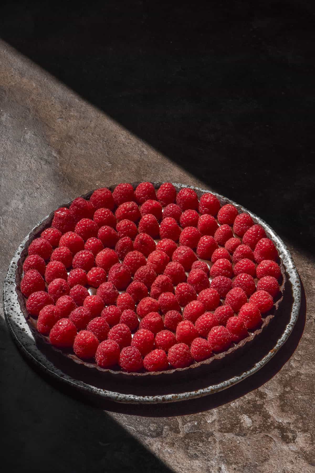 A gluten free chocolate tart covered in raspberries in dramatic contrasting sunlight