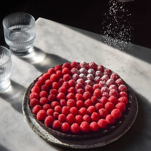 A moody image of a chocolate tart topped with raspberries being sprinkled with icing sugar atop a white marble table in contrasting sunlight