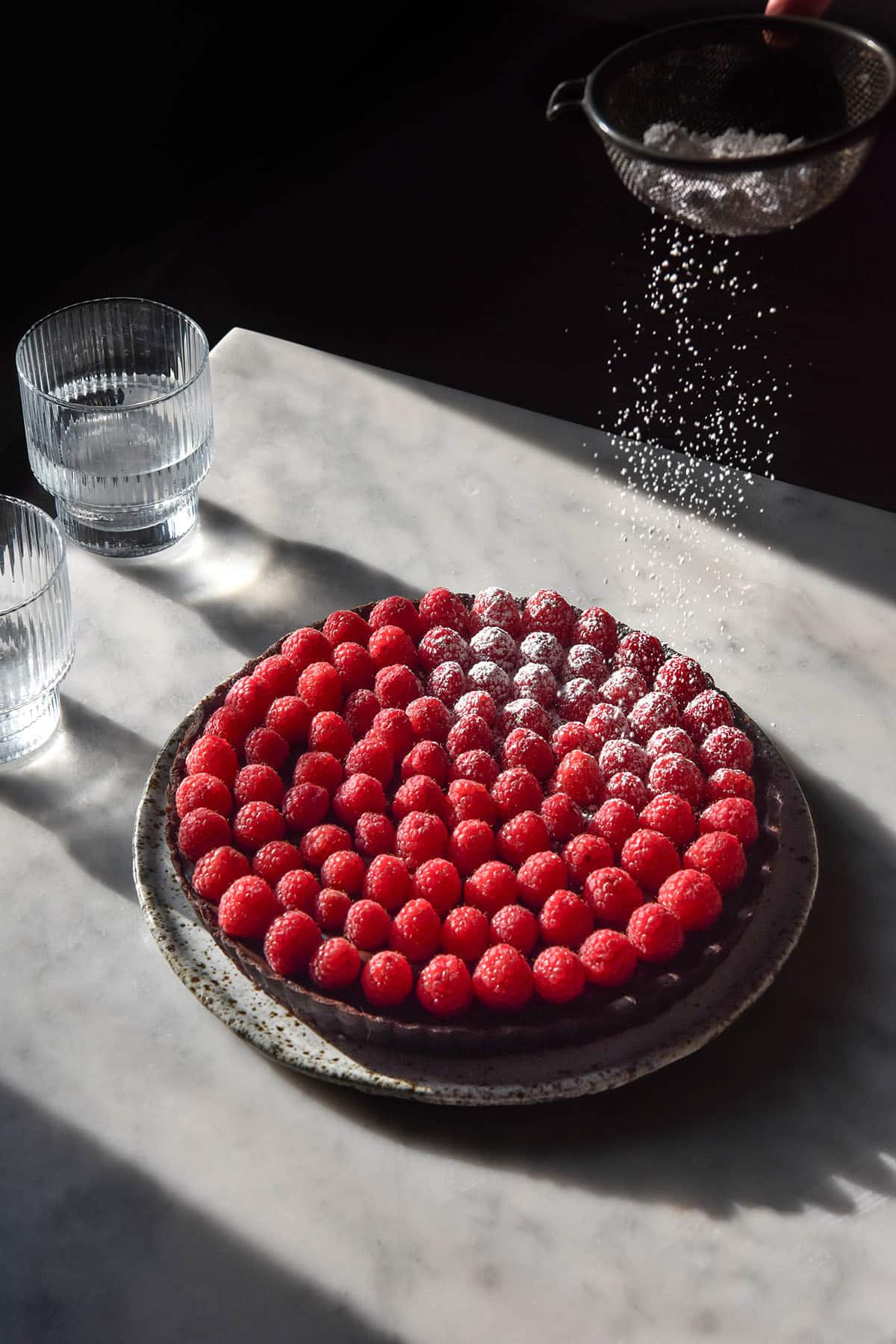 A moody image of a chocolate tart topped with raspberries being sprinkled with icing sugar atop a white marble table in contrasting sunlight