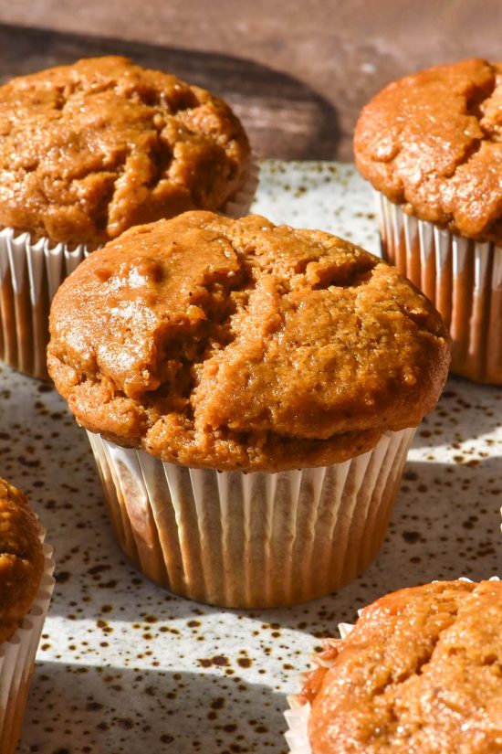 A brightly lit side on image of gluten free vegan pumpkin muffins in white patty pan liners on a white speckled ceramic plate