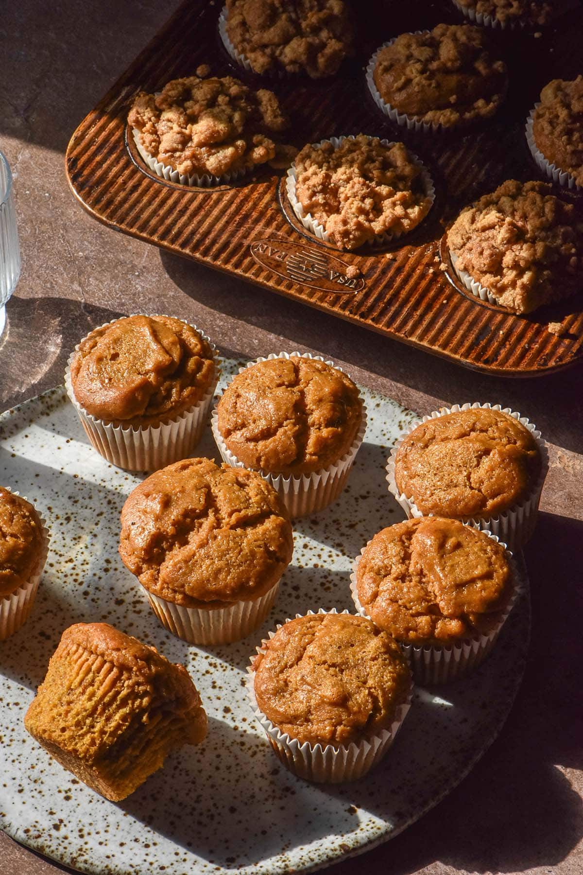 Two batches of gluten free vegan pumpkin muffins on a brown backdrop in contrasting sunlight. One batch of muffins is plain and sits in the foreground on a white speckled ceramic plate. One batch of muffins is topped with streusel and sits in a muffin tray at the back of the image.