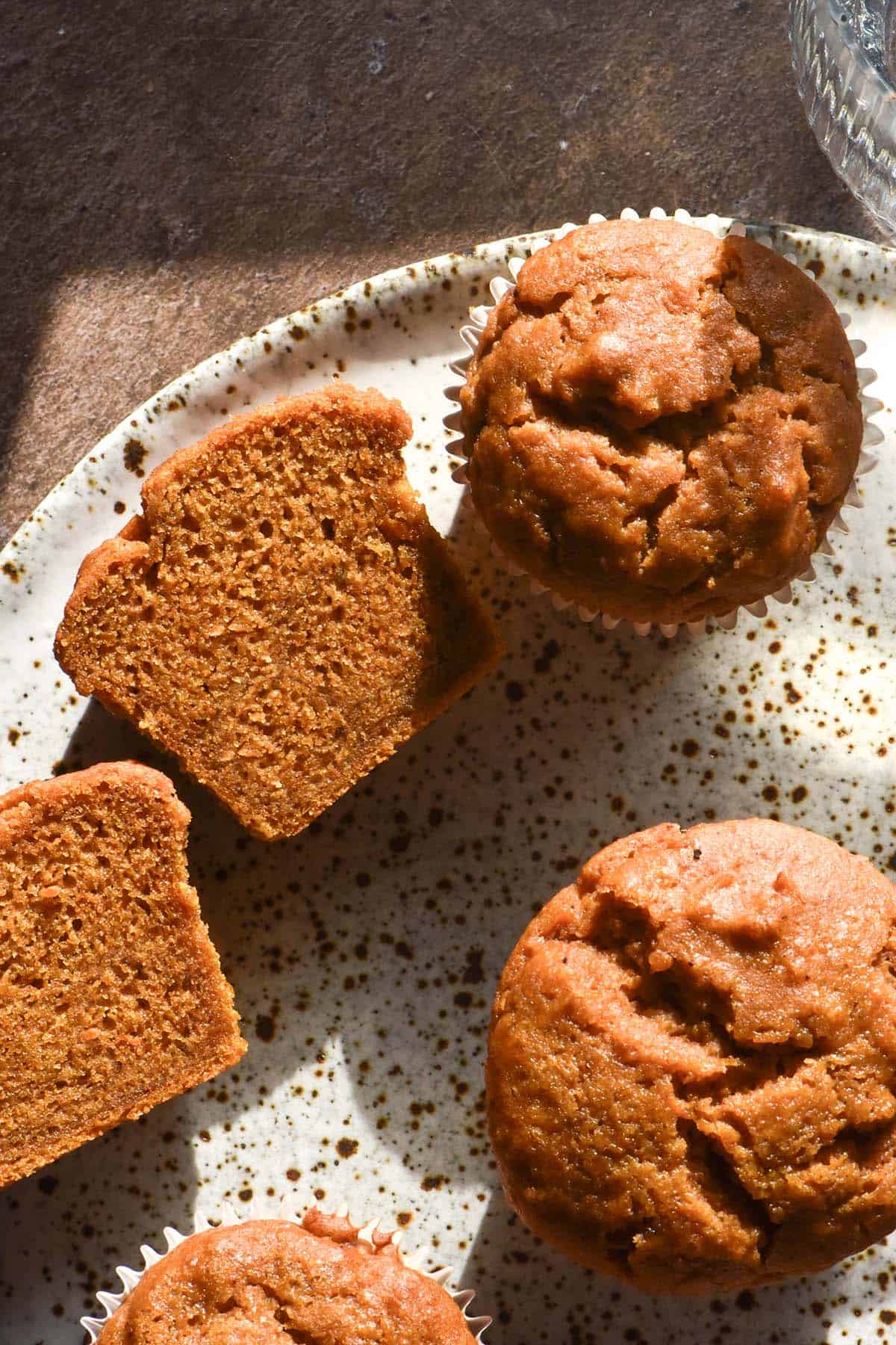 An aerial image of gluten free vegan pumpkin muffins on a white speckled ceramic plate atop a sunlight brown backdrop. One muffin has been sliced open revealing the inner crumb.