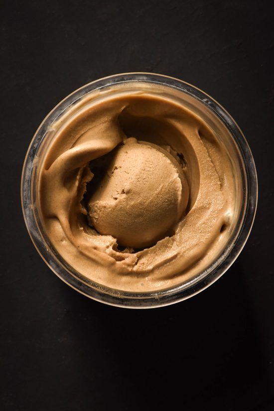 An aerial image of a tub of Ninja Creami vegan gingerbread ice cream on a black backdrop. The ice cream has a scoop in the centre.