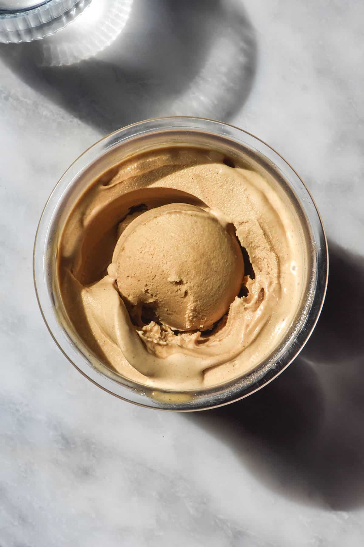 An aerial image of a tub of vegan Ninja Creami gingerbread ice cream on a white marble table in bright sunlight. A sunlit water glass sits in the top left corner.