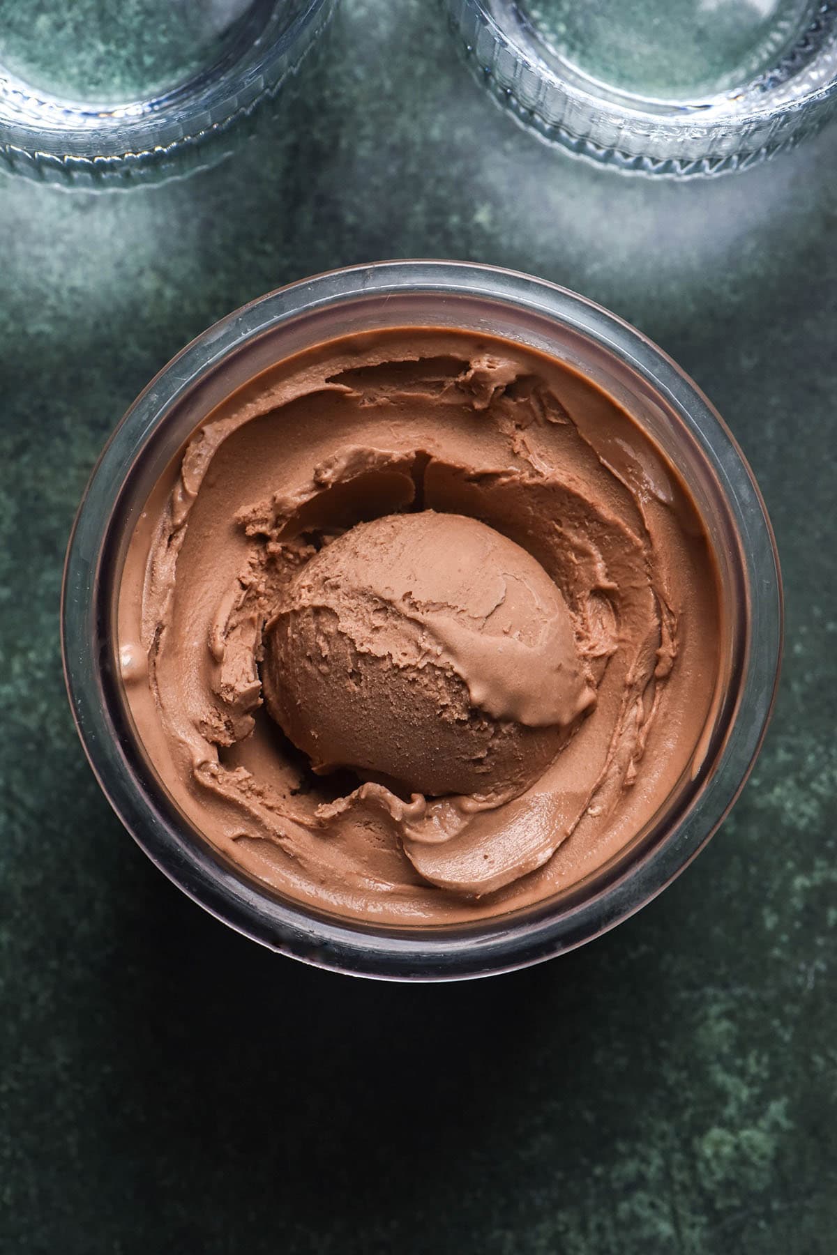 An aerial image of a tub of vegan Ninja Creami Nutella ice cream on an olive green backdrop. Two glasses of water sit in the top of the image.
