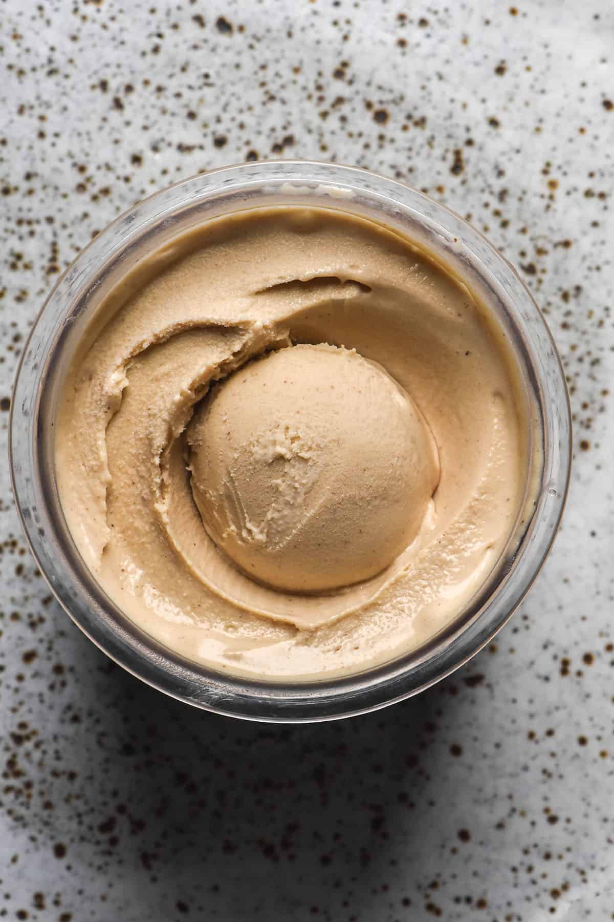An aerial image of a tub of vegan brown butter Ninja Creami ice cream atop a white speckled ceramic plate.