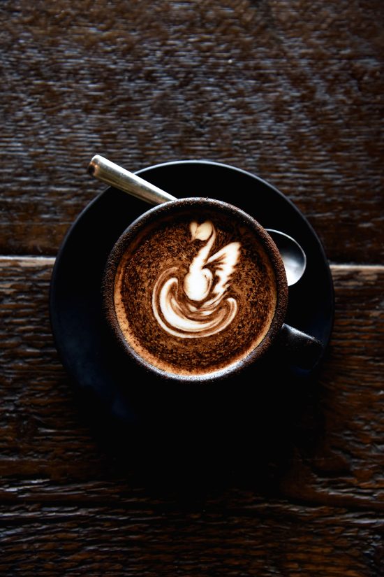 An aerial image of a cappuccino topped with latte art on a black saucer atop a dark wood backdrop.