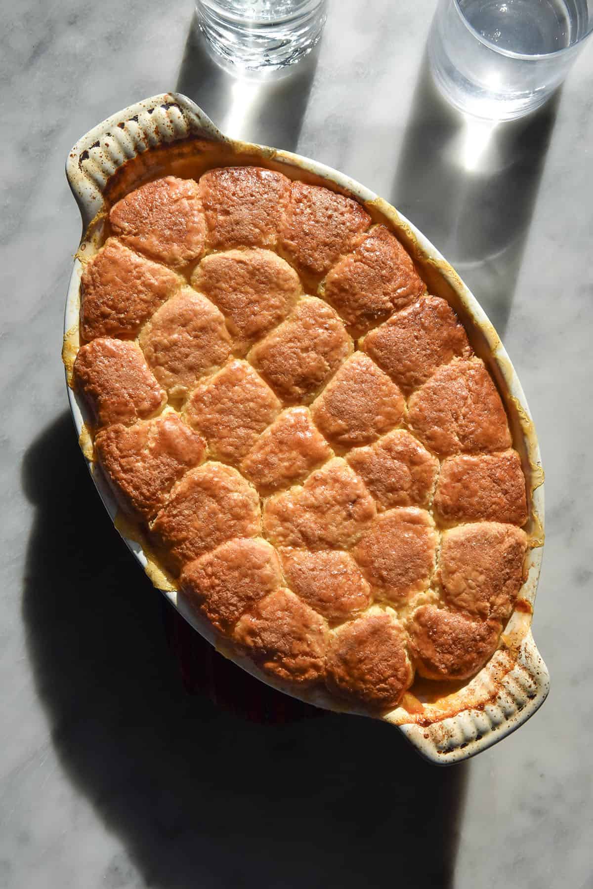 An aerial image of a gluten free peach cobbler in a rectangular baking dish on a white marble table in bright sunlight