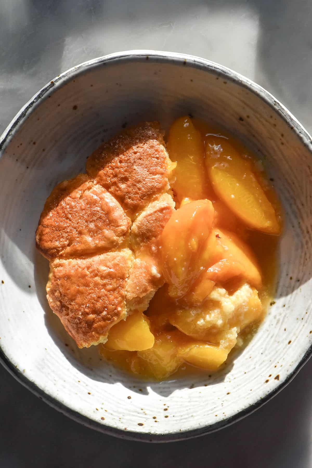 An aerial image of a white speckled ceramic bowl filled with gluten free peach cobbler atop a white marble table in contrasting sunlight
