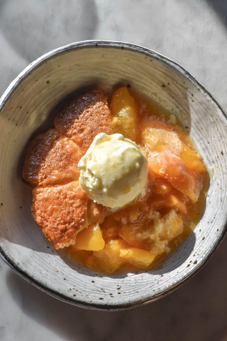 An aerial image of a white speckled ceramic bowl filled with gluten free peach cobbler topped with a scoop of vanilla ice cream. The bowl sits atop a white marble table in contrasting sunlight