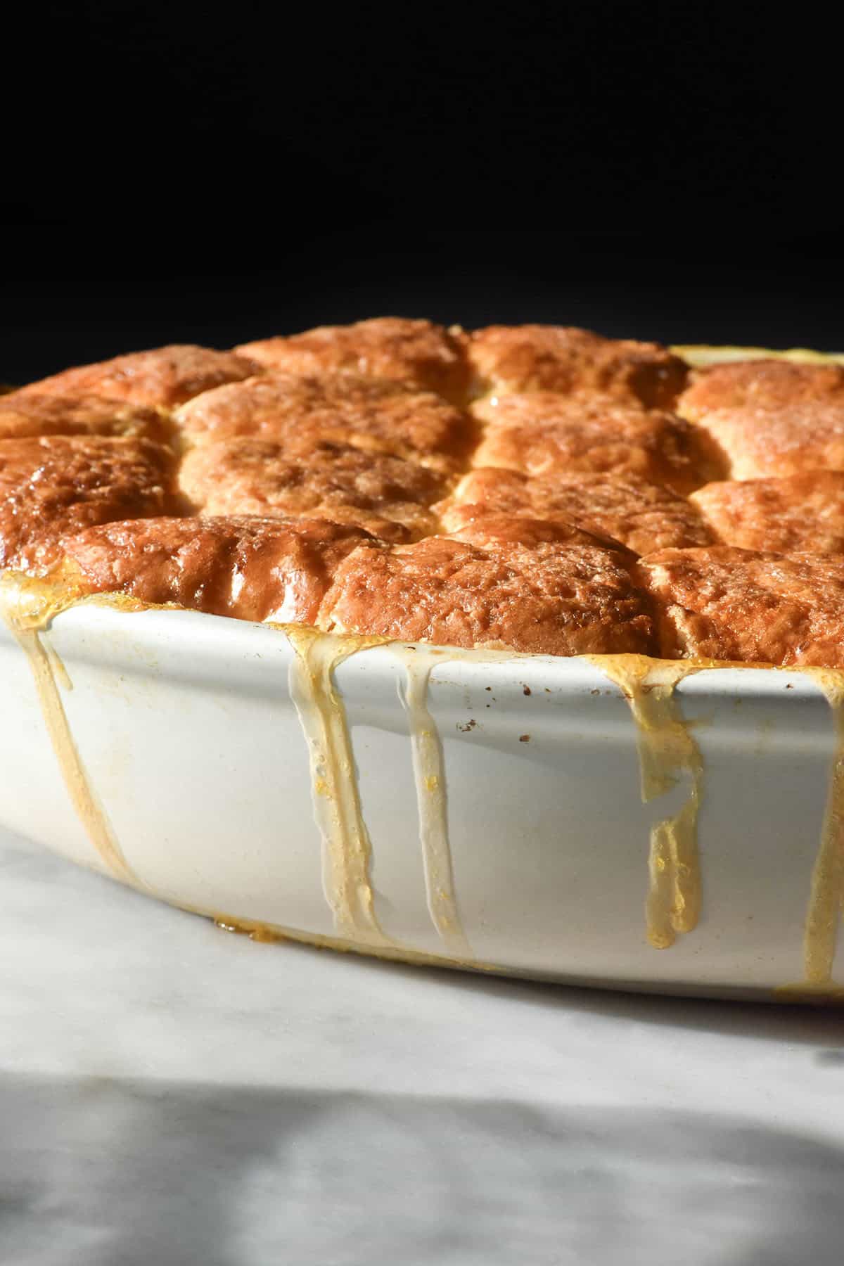 A side on image of a gluten free peach cobbler with juices drizzling down the side of the white baking dish. It sits atop a white marble table against a dark backdrop.