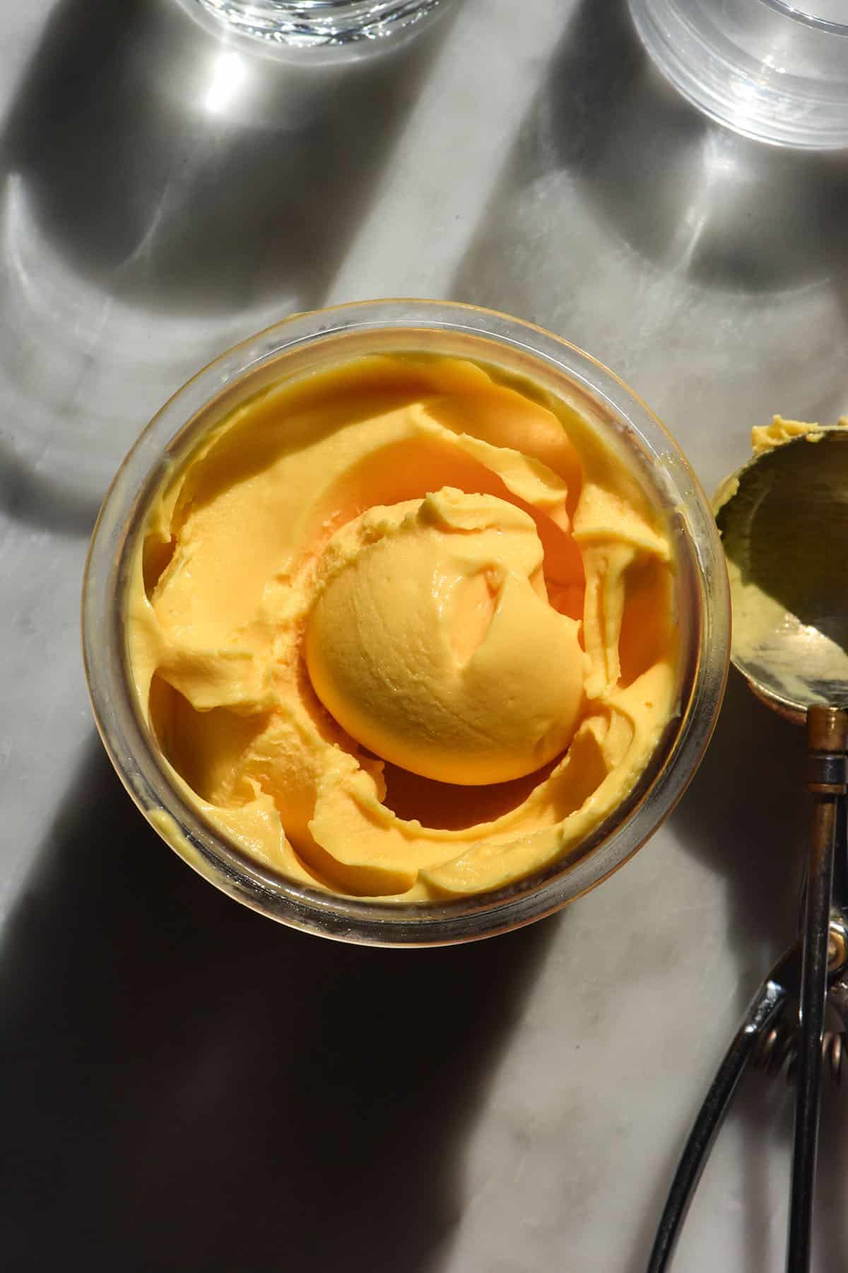 A sunlight aerial image of a tub of Ninja Creami mango ice cream with a scoop in the centre. The ice cream scoop sits to the right of the tub on a white marble table and two sun lit water glasses sit in the top of the image.