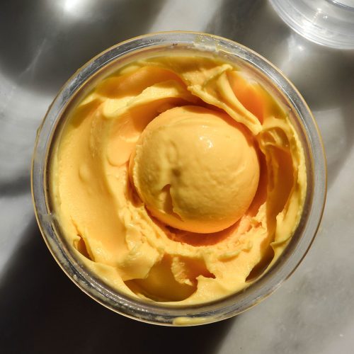 An aerial image of a tub of Ninja Creami mango ice cream on a white marble table in bright sunlight. There is a scoop of mango ice cream in the centre of the tub.