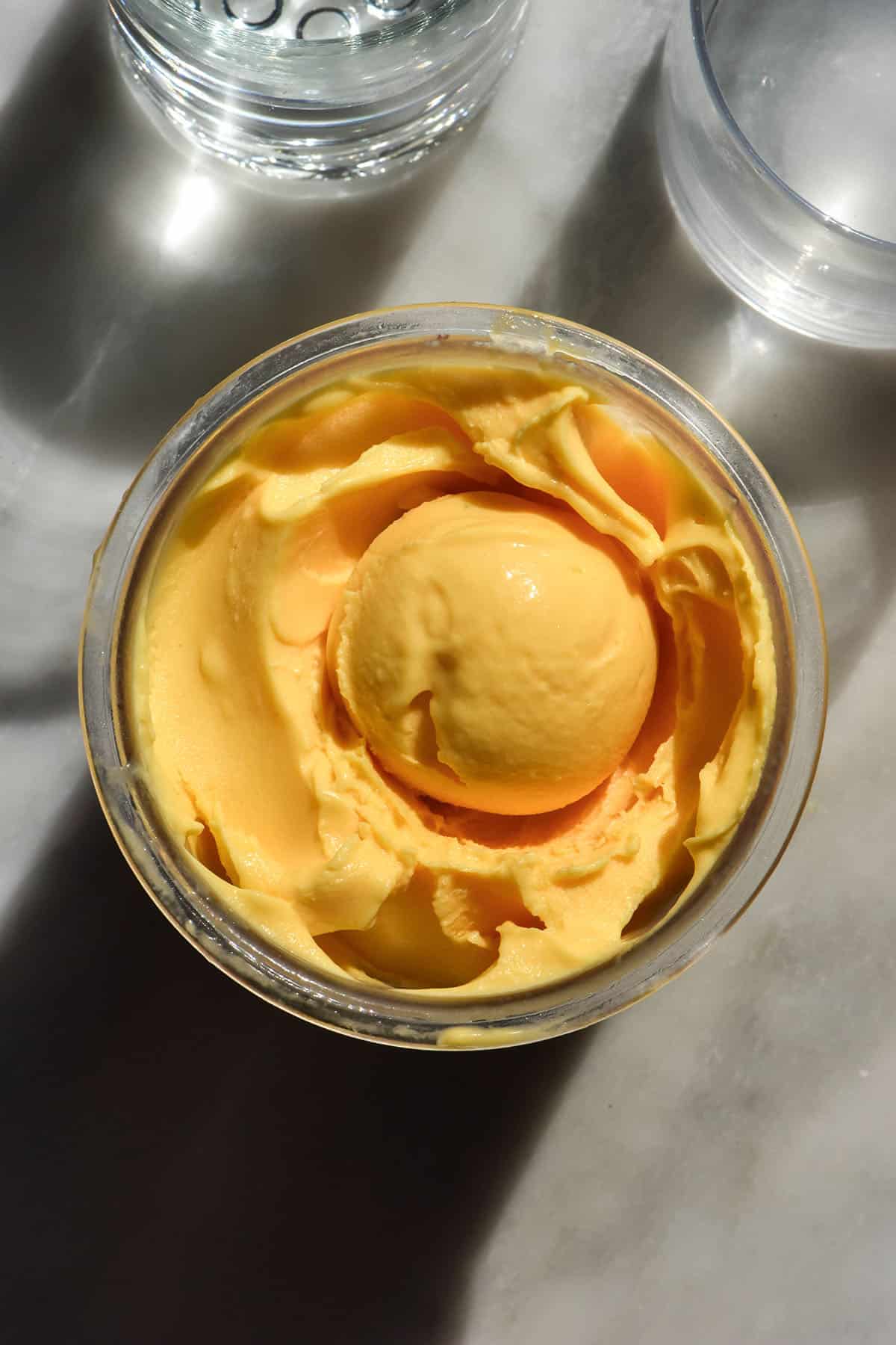 An aerial image of a tub of Ninja Creami mango ice cream on a white marble table in bright sunlight. There is a scoop of mango ice cream in the centre of the tub.