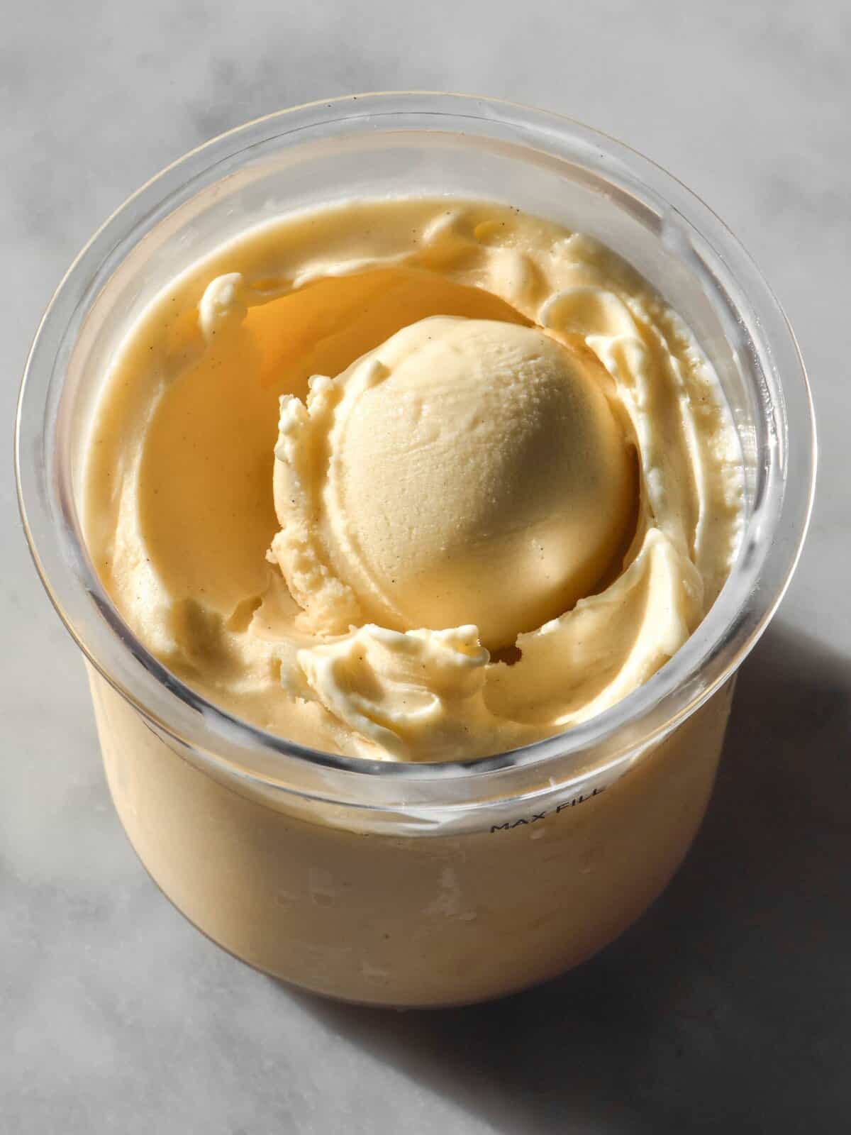An aerial image of a tub of Ninja Creami custard ice cream atop a brightly lit white marble table. The ice cream has a scoop in the centre.