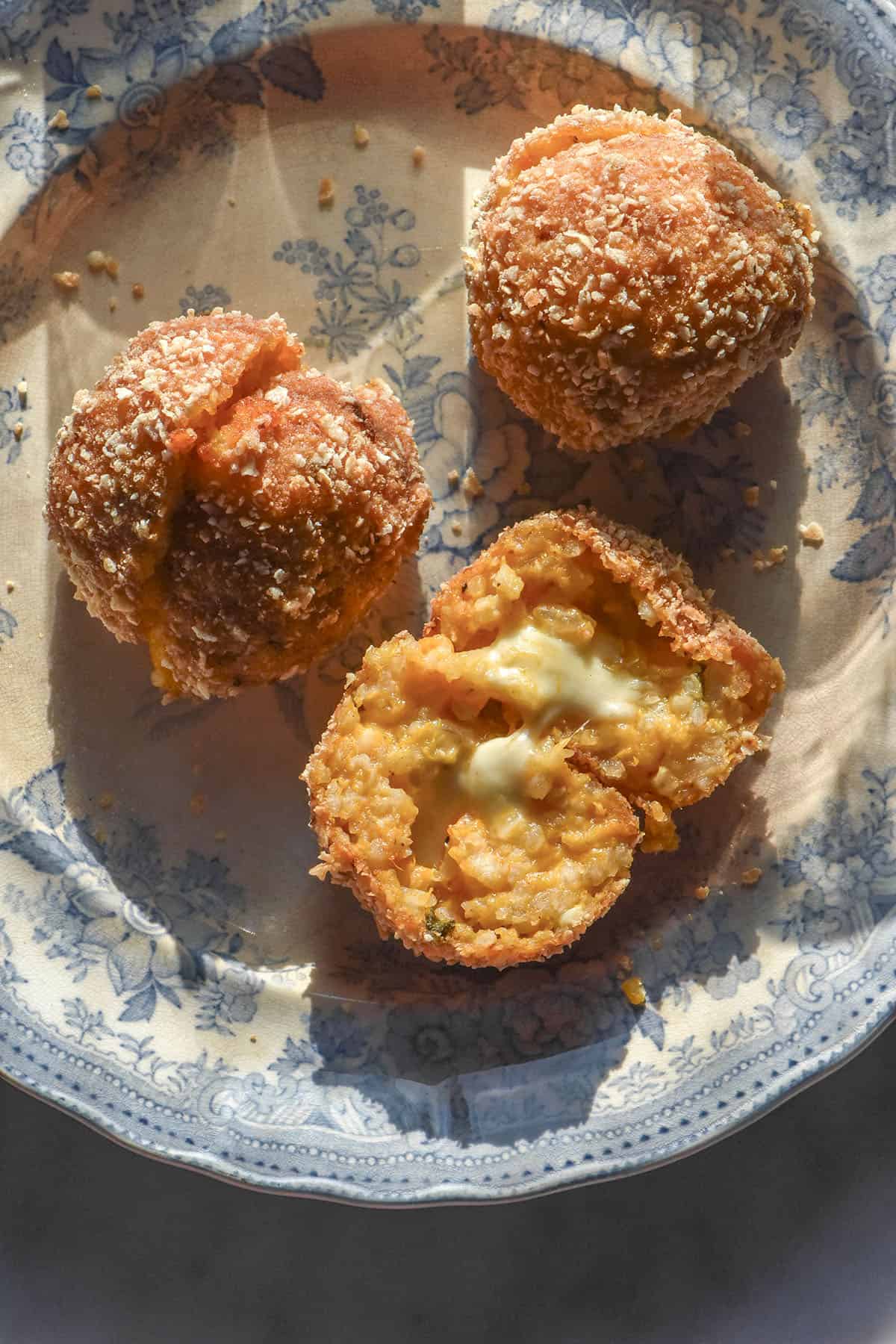 An aerial image of gluten free arancini that have been breaded with gluten free panko crumbs. The arancini sit atop a vintage blue and white plate in contrasting sunlight.