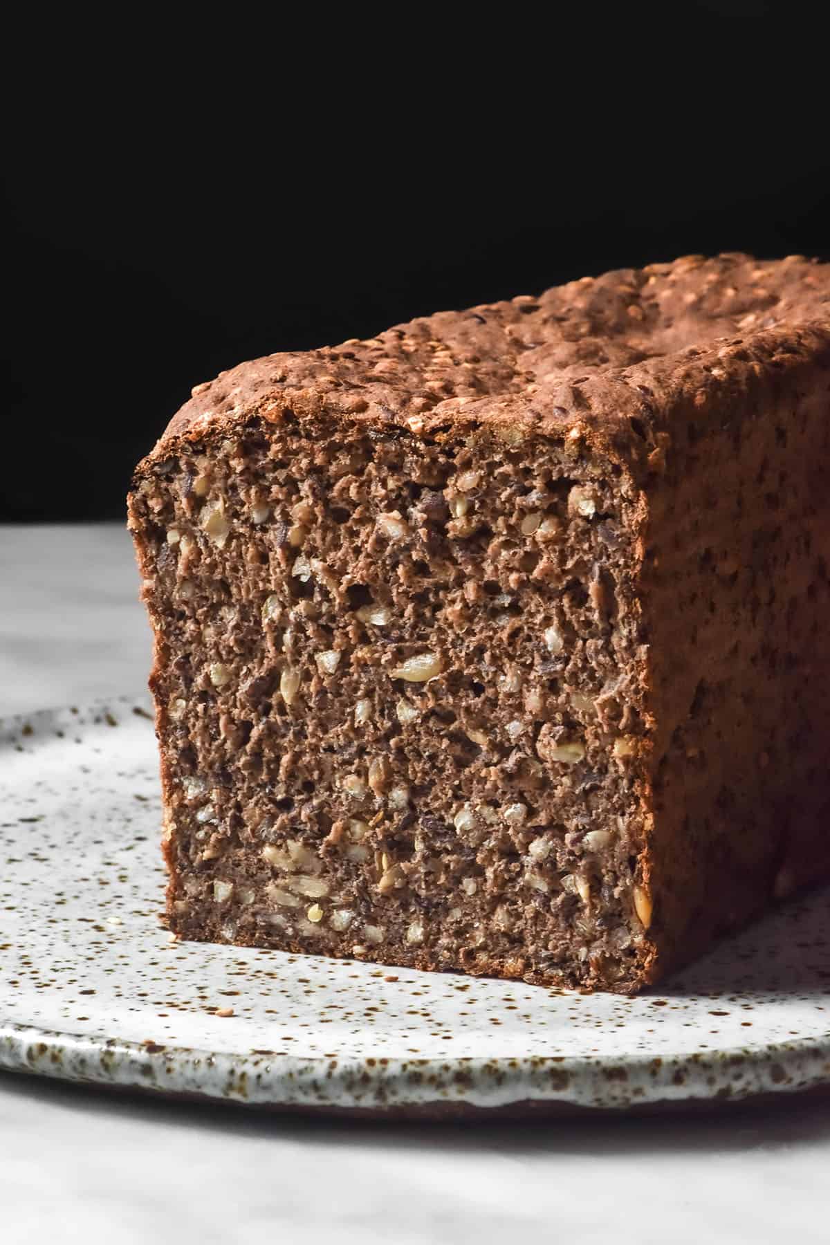 A side on image of a loaf of gluten free seeded rye bread on a white speckled ceramic plate against a dark backdrop. The loaf has been sliced to reveal the seeded and dense inner crumb.