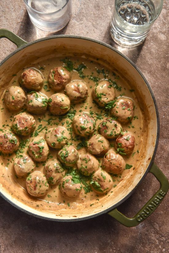 An aerial image of a skillet filled with tofu meatballs in a cream sauce and topped with chopped parsley. The skillet sits atop a light brown backdrop and two water glasses sit in the top of the image.