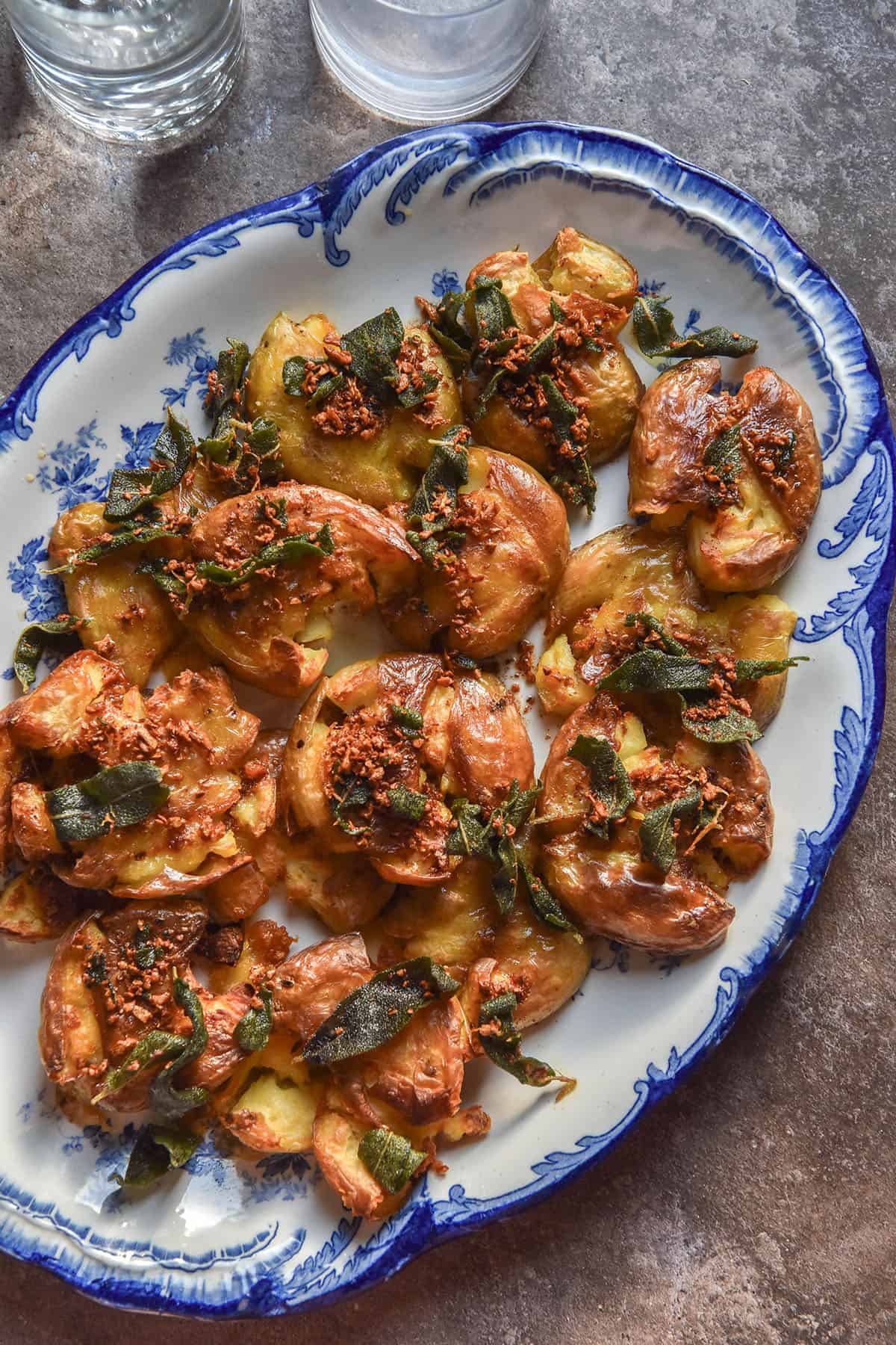 An aerial image of crispy brown butter, sage and garlic potatoes on a blue and white serving plate atop a brown backdrop. Two sunlit water glasses sit in the top left of the image.