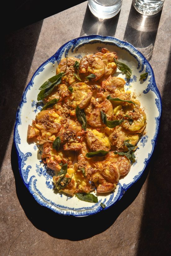 An aerial sunlit image of low FODMAP garlic sage potatoes on a blue and white serving platter atop a dark brown backdrop. Two sunlit glasses of water sit in the top right of the image.