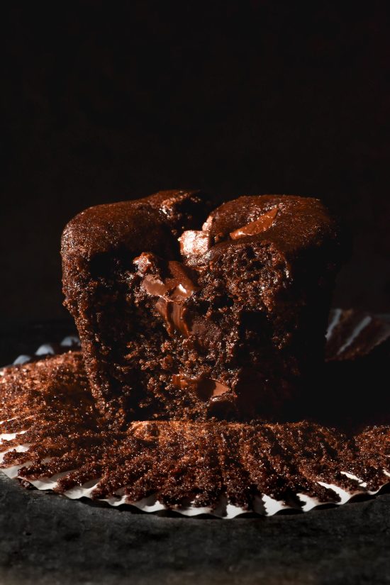 A macro close up image of a chocolate buckwheat muffin sitting atop a muffin liner against a dark backdrop. The muffin has been bitten into, revealing the inner crumb and chocolate chips inside.