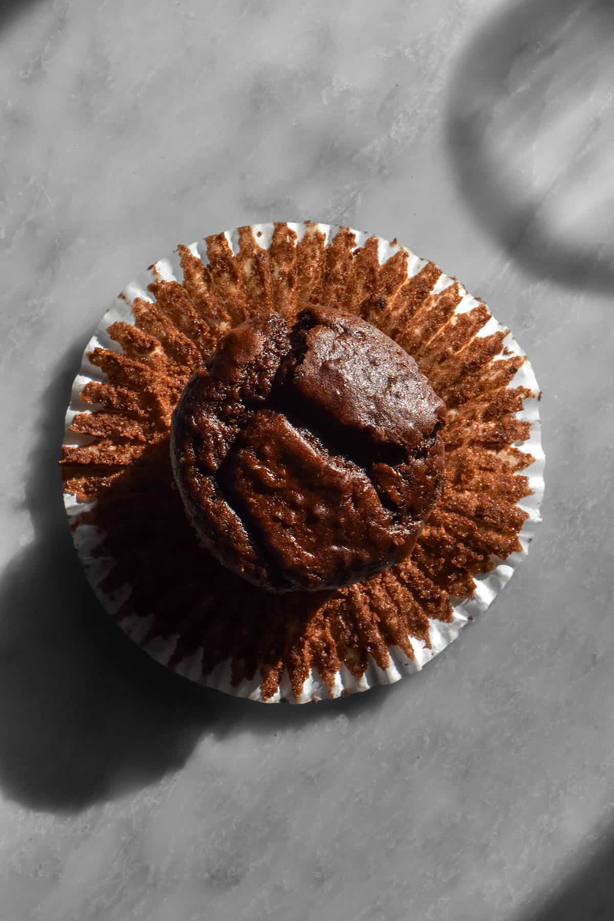 An aerial sunlit image of a chocolate buckwheat muffin sitting atop a muffin liner on a white marble table. A sunlit glass of water sits to the top right of the image