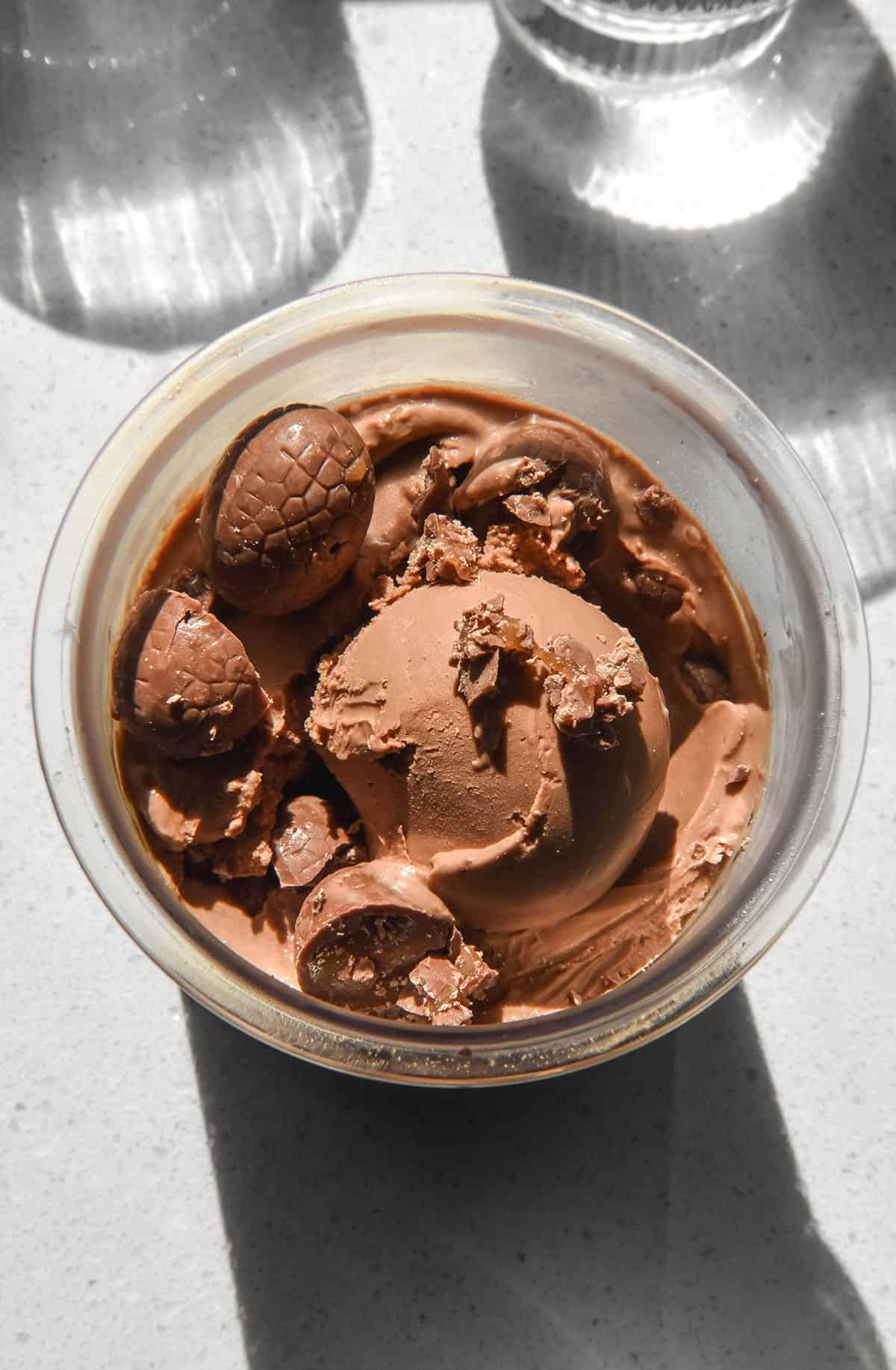 An aerial sunlit image of a tub of Ninja Creami chocolate caramel ice cream topped with chocolate caramel Easter eggs on a sunlit backdrop.