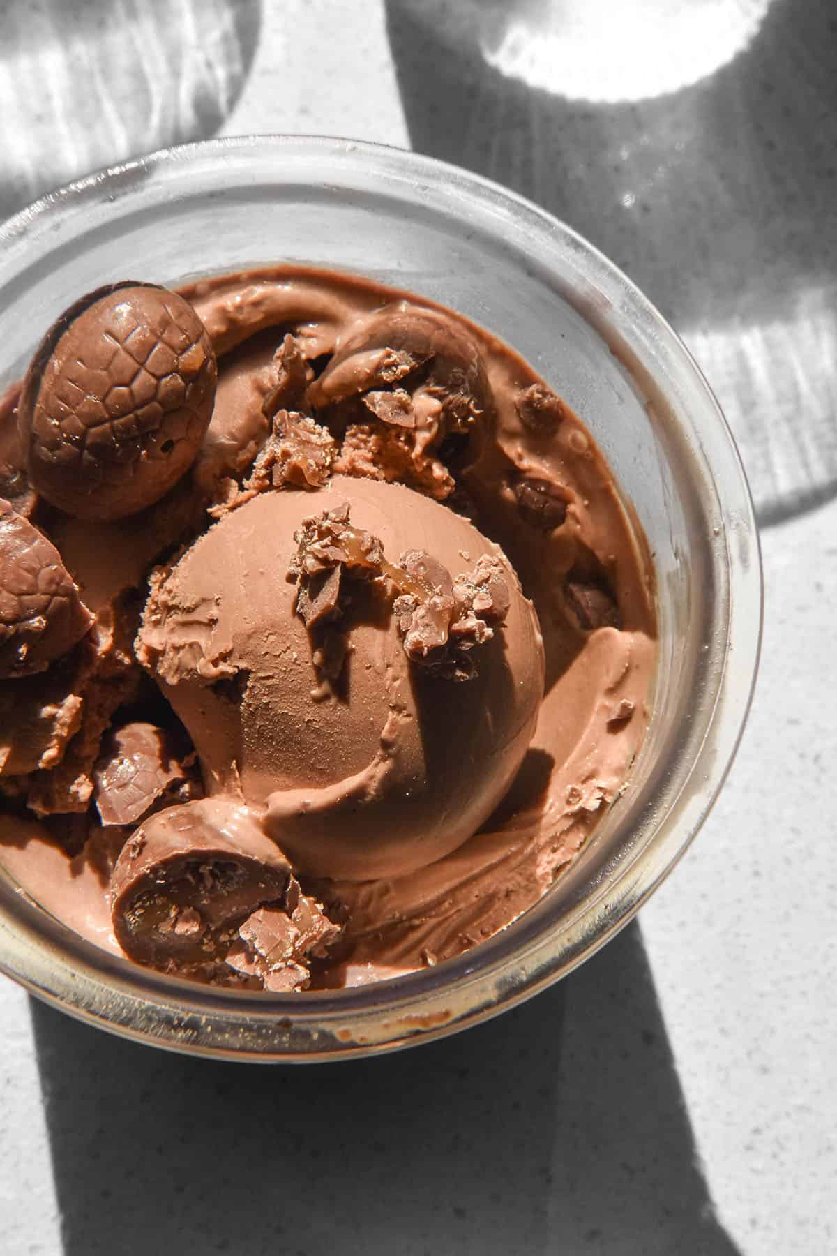An aerial sunlit image of a tub of Ninja Creami chocolate caramel ice cream topped with chocolate caramel Easter eggs on a sunlit white backdrop.