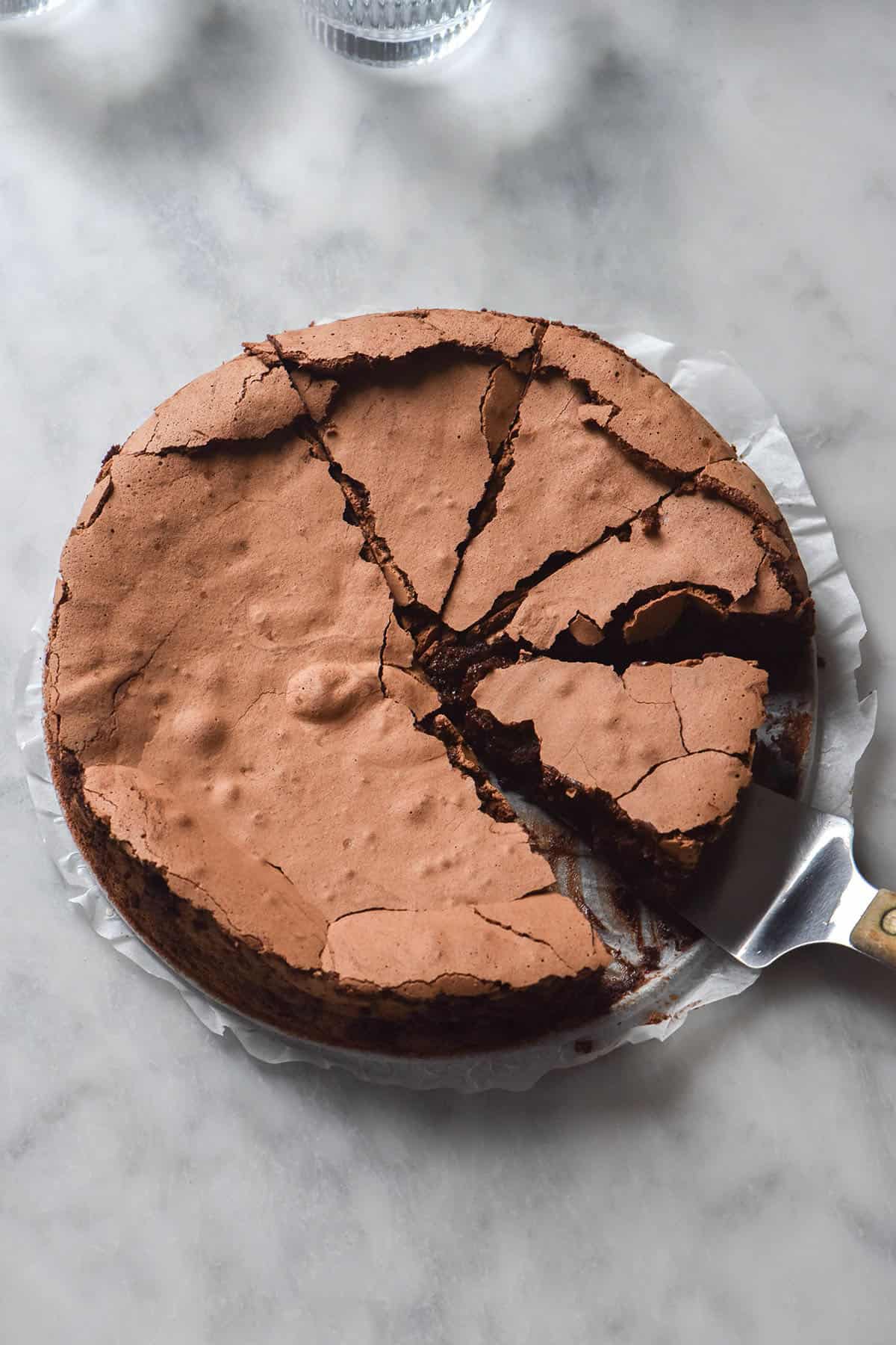 An aerial image of a dairy free flourless chocolate cake on a white marble table with water glasses in the top left of the image. A cake slice sits on a cake server with a wooden handle.