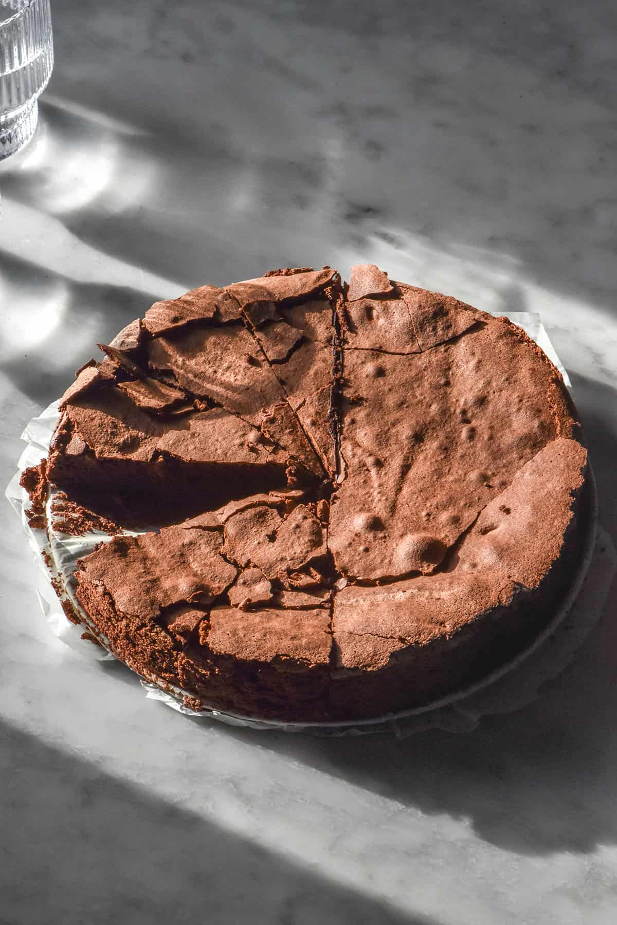 An aerial image of a dairy free flourless chocolate cake on a sunlit white marble table with sunlit water glasses in the background. The cake has been slice and one slice has been removed.