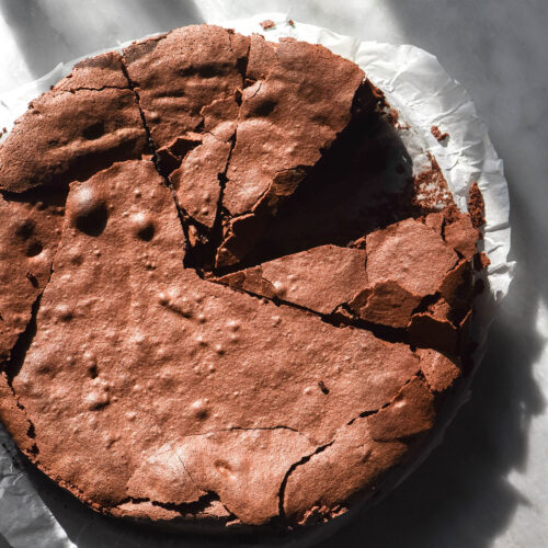 An aerial image of a dairy free flourless chocolate cake on a sunlit white marble table with sunlit water glasses in the background. The cake has been slice and one slice has been removed.