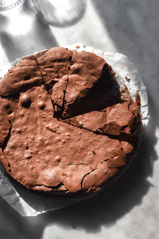 An aerial image of a dairy free flourless chocolate cake on a sunlit white marble table with sunlit water glasses in the background. The cake has been slice and one slice has been removed.