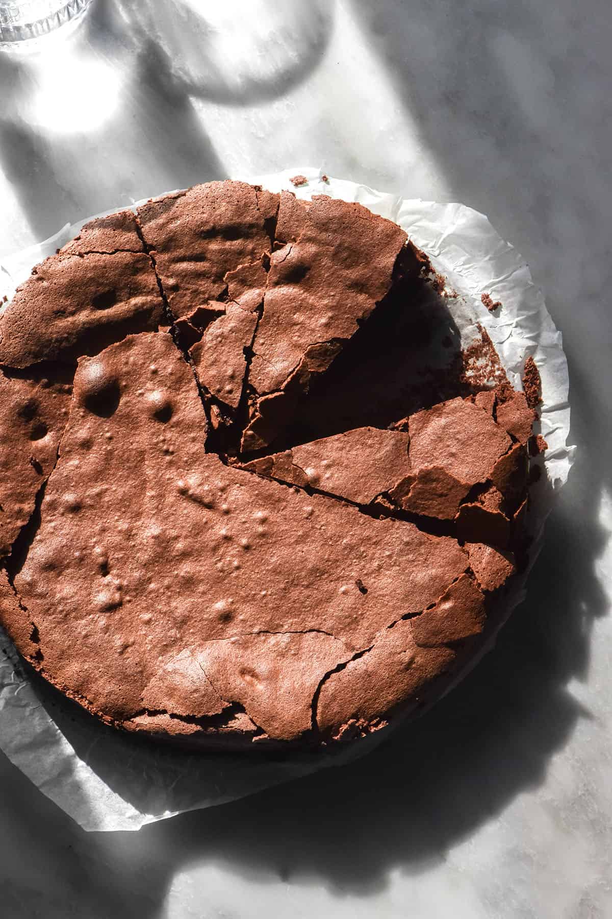 An aerial image of a dairy free flourless chocolate cake on a sunlit white marble table with sunlit water glasses in the background. The cake has been slice and one slice has been removed.