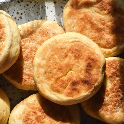 A sunlit aerial image of gluten free English muffins in a stack on a white ceramic plate against a white marble backdrop