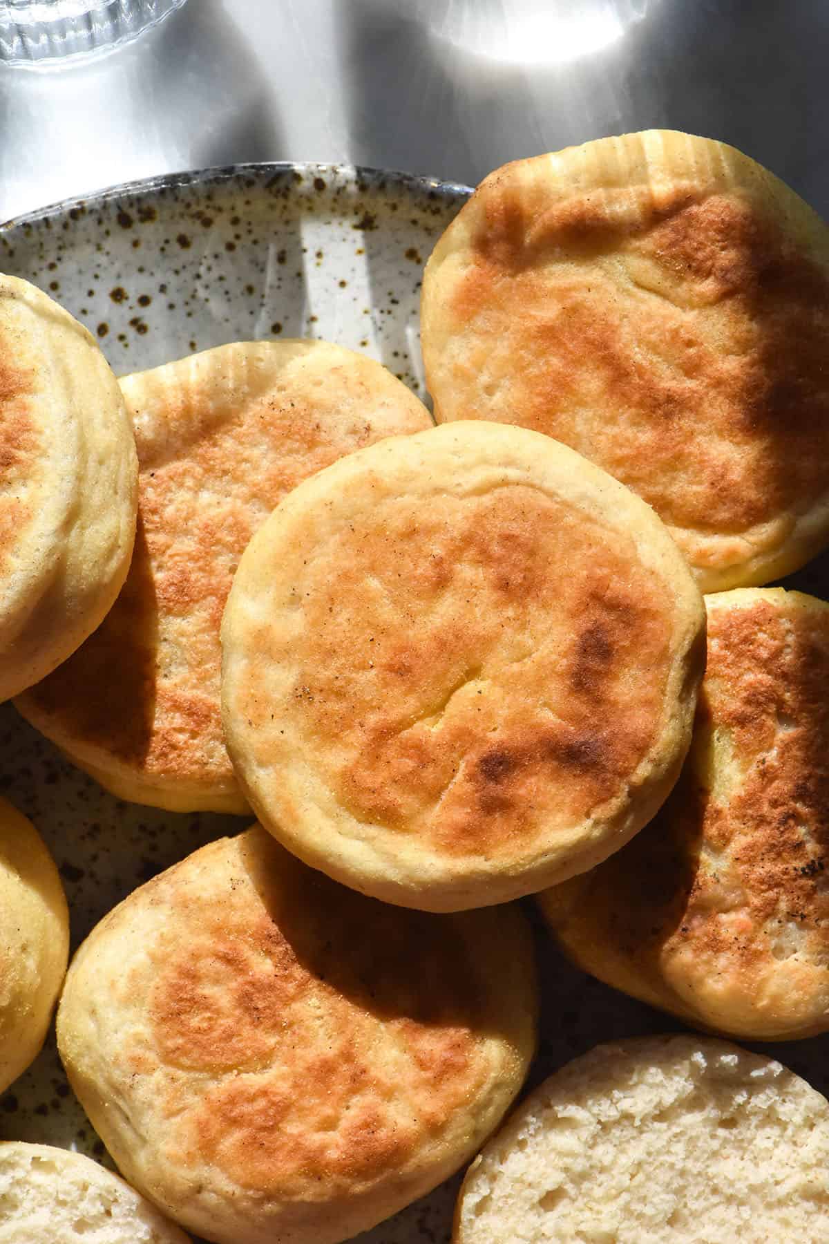 A sunlit aerial image of gluten free English muffins in a stack on a white ceramic plate against a white marble backdrop