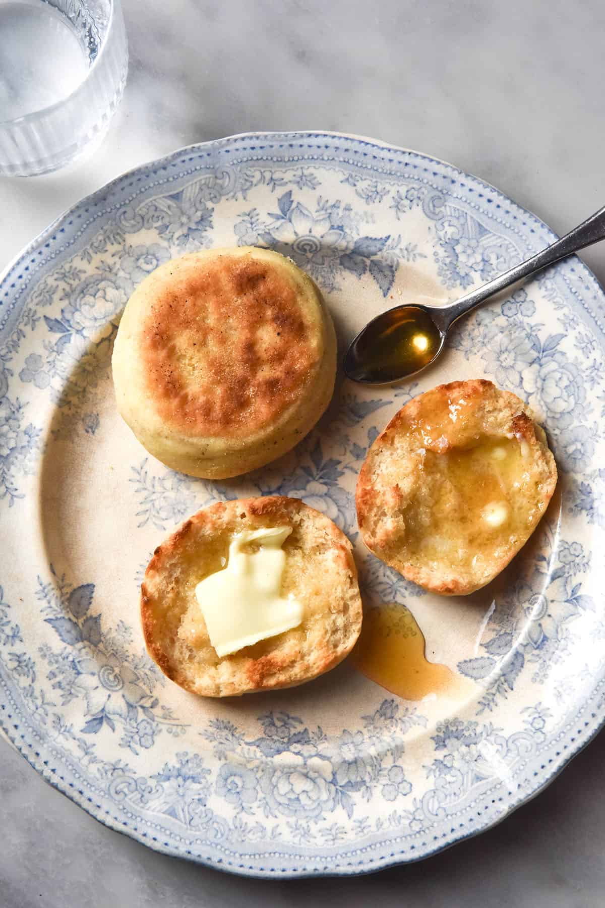 An aerial image of a white and blue ceramic plate topped with two gluten free English Muffins, one of which is toasted and smothered in butter and honey. The plate sits atop a white marble table and a sunlit glass of water sits to the top left of the image.