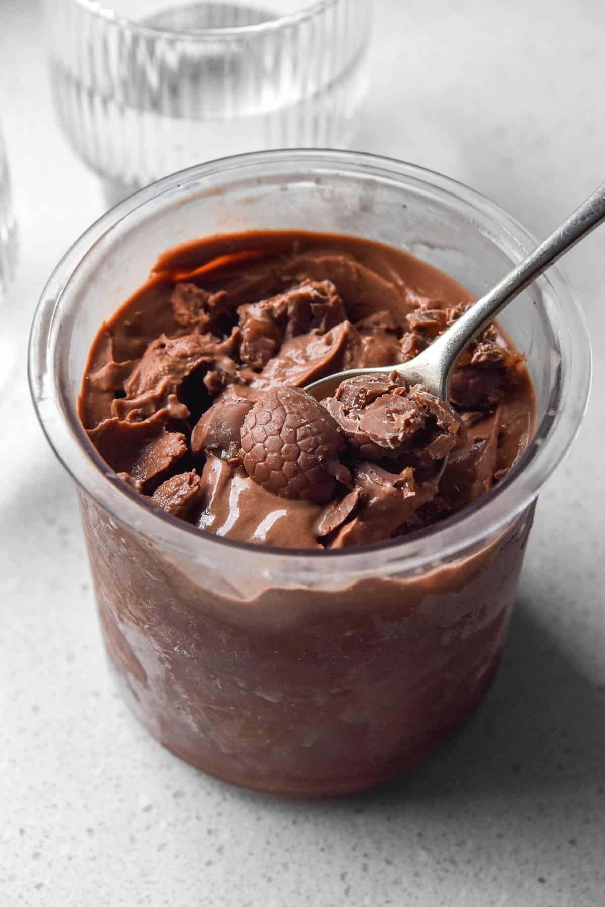 A sunlit image of a tub of Ninja Creami chocolate caramel ice cream on a white stone backdrop. The ice cream has been topped with chocolate caramel Easter eggs and a spoon sticks out of the right side of the tub.