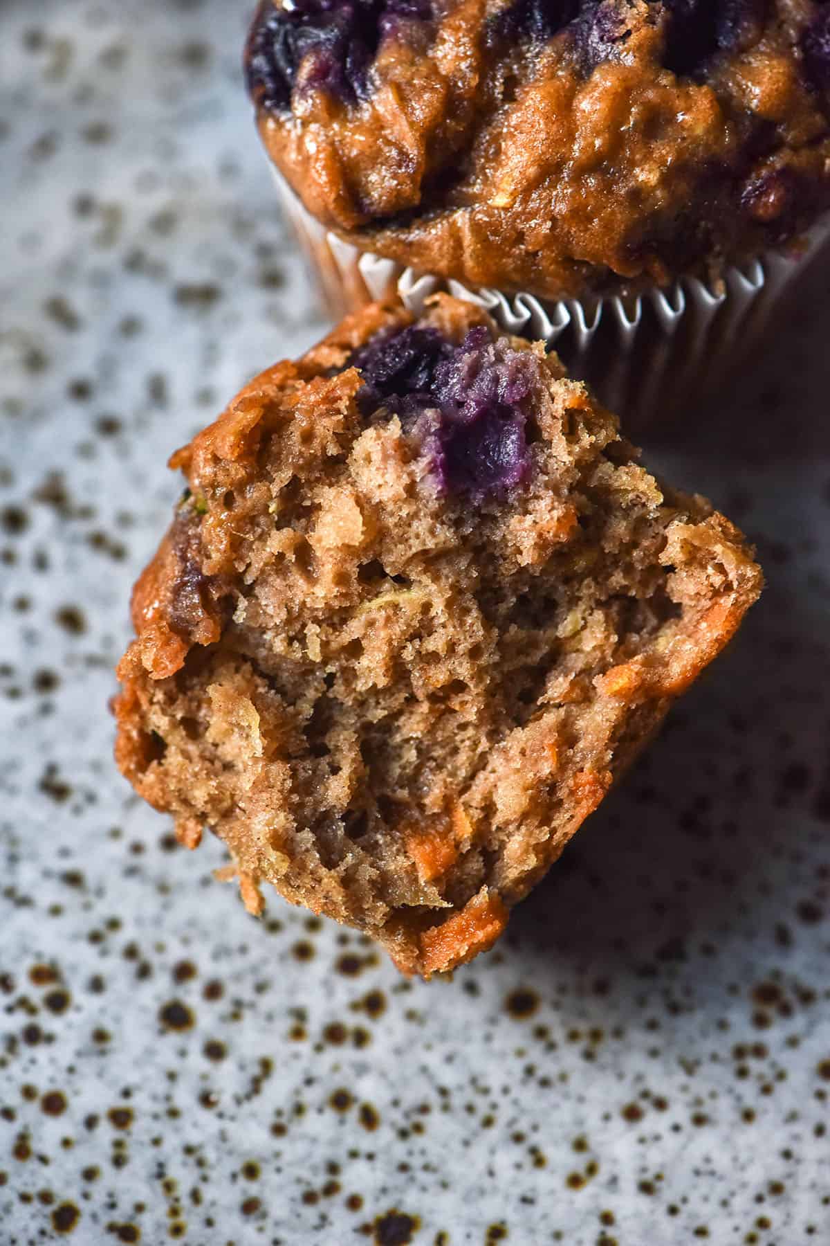 An aerial image of a gluten free buckwheat breakfast muffin that has been halved to reveal the soft inner crumb. The muffin is studded with blueberries and shredded carrot, zucchini and apple and sits on a white speckled ceramic plate.