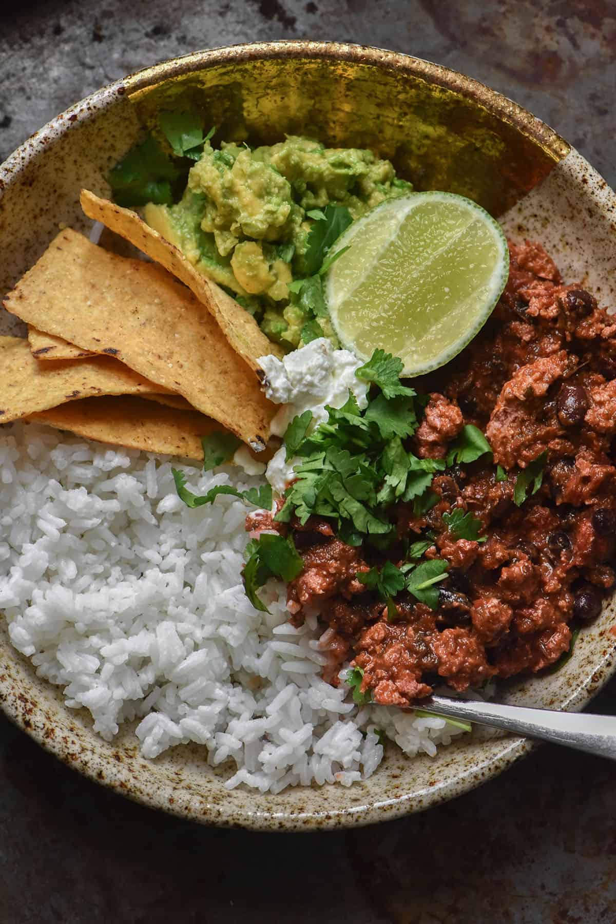 An aerial image of a speckled ceramic bowl filled with rice, low FODMAP vegan nomato chilli, guacamole, corn chips and a wedge of lime. The bowl sits on a speckled steel backdrop.